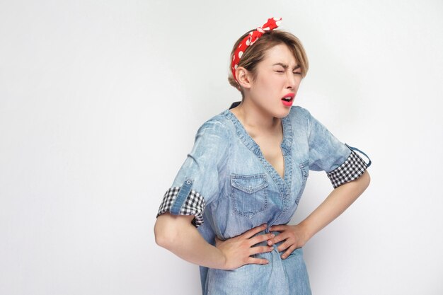 Stomach pain or period. Portrait of beautiful young woman in casual blue denim shirt with makeup, red headband standing and holding her painful belly. indoor studio shot, isolated on white background.