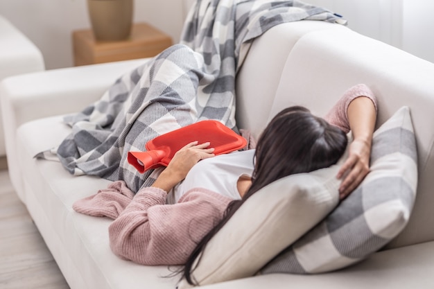 Stomachache easing by thermophore while woman lies on a couch under the blanket.