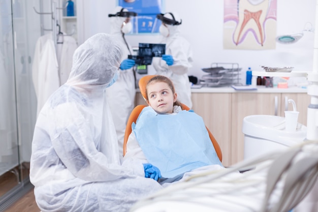 Stomatologist during covid19 wearing ppe suit doing teeth procedure of child sitting on chair.