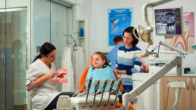 Stomatologist holding plaster model of the mandible speaking with girl patient. Dentist showing the correct dental hygiene using mock-up of skeleton of teeth, sample of human jaw with toothbrush