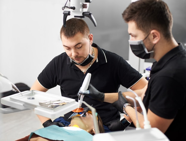 Stomatologist looks at the patient while his assistant is completing the process of filling the tooth in modern clinic. Both are dressed in black uniforms, masks and gloves