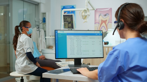 Infermiera stomatologa che parla con i pazienti utilizzando l'auricolare che fa appuntamenti dal dentista seduto di fronte al computer mentre il medico sta lavorando con lo sfondo del paziente esaminando il problema dei denti