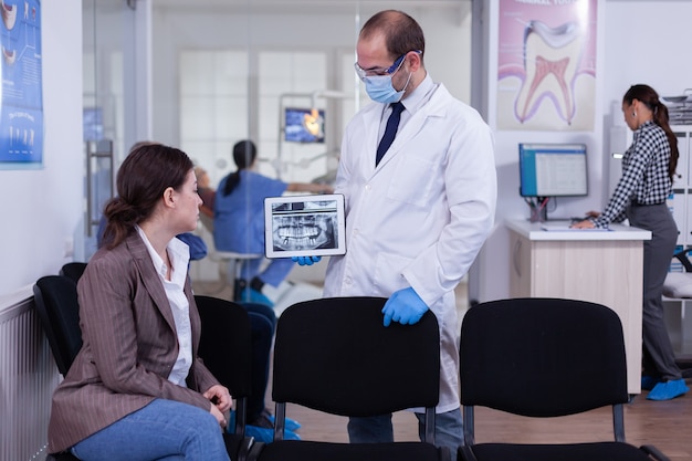 口腔病学クリニックの待合室で椅子に座っている女性にX線を説明するデジタル画面を指している口腔病学者
