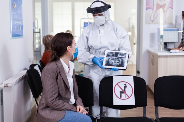 Stomatologist in protective suit pointing on digital x-ray of tooth explaining to patient treatment using tablet in covid-19 pandemic