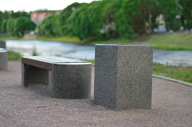 A stone bench and a garbage can next to a paving stone path in a quiet city park on a sunny summer day against the background of trees