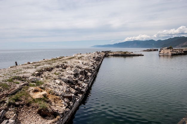 Каменные курганы старого разрушенного пирса на Байкале. Вода спокойная. Холмы в дымке на горизонте. Пасмурно. По горизонтали.