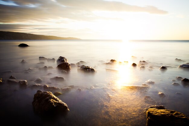 Photo stones in ocean against sunset, over which fog. sky is filled with clouds. far beyond horizon there mountains