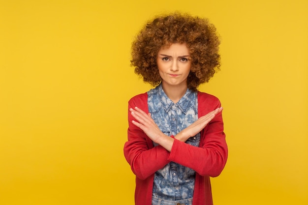 Stop, banned access. Portrait of young curly-haired woman crossing hands, warning with x sign, rejecting troubles with definitive no, body language. indoor studio shot isolated on yellow background