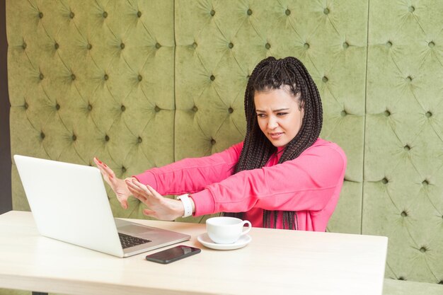 Stop I dont want to look at this.Portrait of scared young girl freelancer with black dreadlocks hairstyle in pink blouse sitting, looking and showing stop at laptop screen and stressed face. Indoor