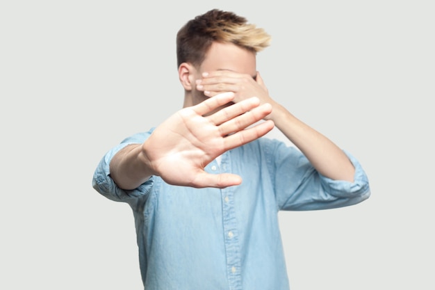 Stop, I don't want to see at this. Portrait of scared handsome young man in light blue shirt standing, covering his face and showing stop hand gesture. indoor studio shot on grey background copy space