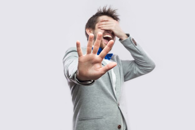 Stop, i don't want to see this. Portrait of shocked or scared young bearded man in casual grey suit standing closed eyes and blocking with hand. indoor studio shot, isolated on light grey background.