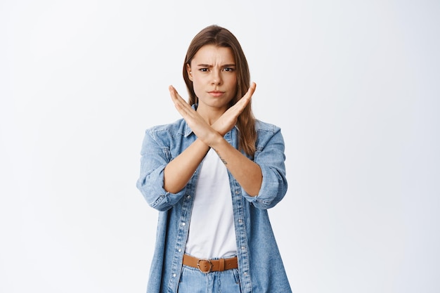 Stop it. Disappointed frowning woman making cross gesture and condemn bad thing, blocking or saying no, standing on white