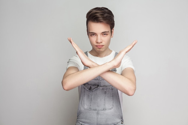 Stop. Portrait of serious young brunette man in casual style with t-shirt and denim overalls standing, looking at camera and showing close way gesture. indoor studio shot, isolated on gray background.