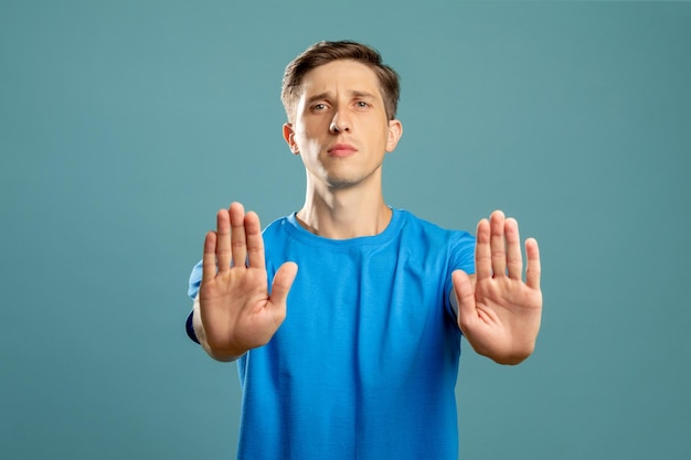 Stop sign refusal gesture forbidden way warning notice\
confident young man holding hands open palms looking at camera\
isolated on blue