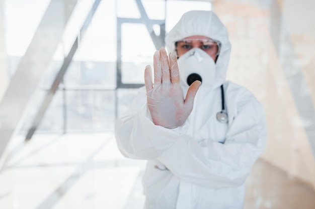 사진 바이러스 제스처를 멈추십시오 실험실 코트 방어 안경과 마스크를 입은 여성 의사 과학자가 실내에 서 있습니다.