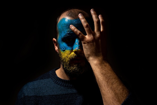 Stop war conflict between ukraine and russia portrait of a
young man with his face painted in the blue and yellow colors of
the flag very sad with a hand on his face