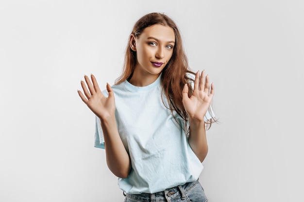 Stop. The woman raises her hands to the camera. The model feels confused and does not know what to choose. Beautiful sweet brunette girl with fashion makeup purple lips on an isolated gray background