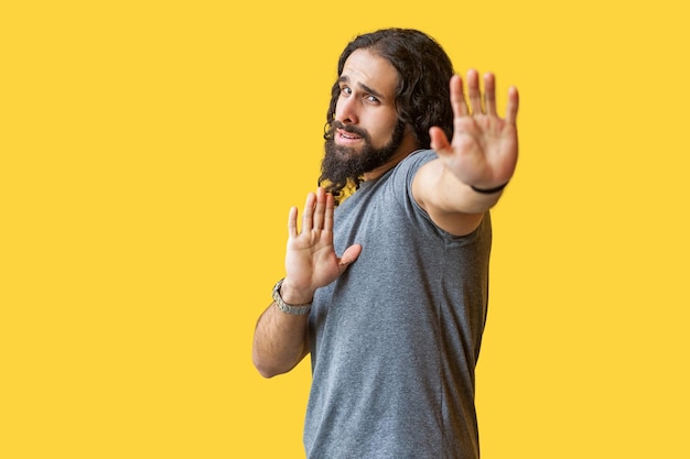 Stoppen. Portret van een bange of verwarde bebaarde jongeman met lang krullend haar in een grijze tshirt die staat met een stop-handtekengebaar en naar de camera kijkt. indoor studio opname geïsoleerd op gele achtergrond.