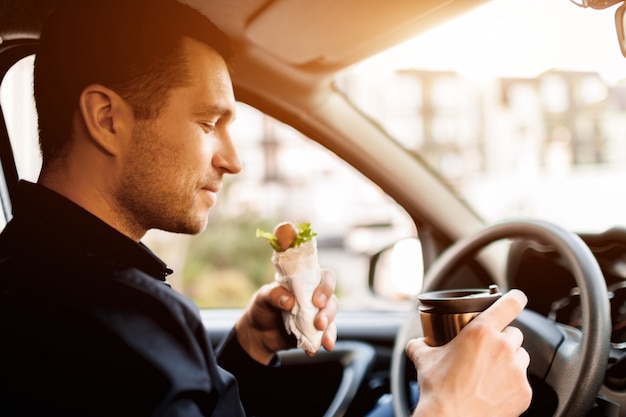 Stoppen voor een hapje. Man eet snack in de auto en drinkt koffie of thee.