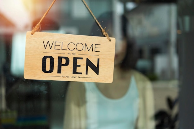 Il proprietario del negozio gira il cartello aperto attraverso il vetro della porta e pronto per il servizio cartello aperto appeso davanti al bar con sfondo astratto chiaro bokeh colorato