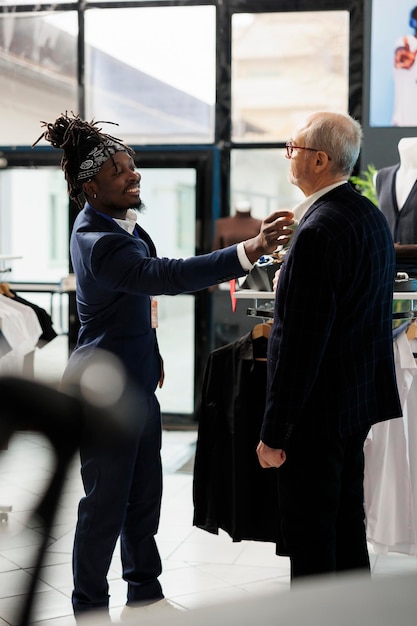 store worker helping stylish customer choosing formal suit in showroom, african american employee helping client to buy trendy clothes. senior man buying fashionable casual wear in modern boutique