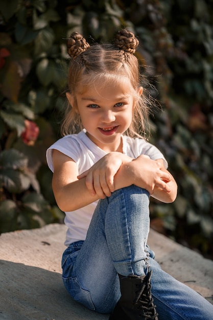 Straatfoto van een klein mooi meisje in jeans en een wit t-shirt op een achtergrond van betonnen platen en herfstbladeren