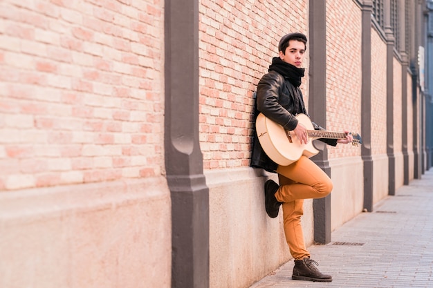 Straatmuzikant die akoestische gitaar speelt jonge knappe man met jas en hoed buitenshuis