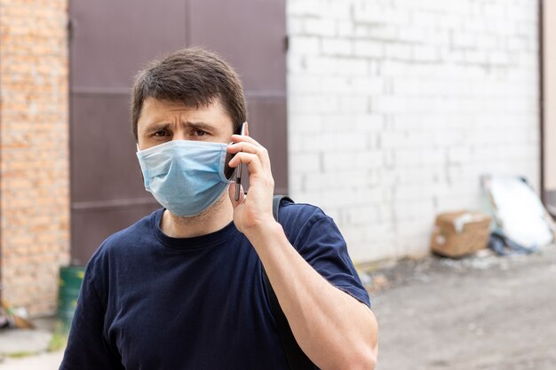 Straatportret van een blanke man die op een smartphone praat in een beschermend blauw medisch masker in de periode van pandemisch coronavirus covid-19. Mannetje in een wegwerpmasker ter bescherming tegen virussen