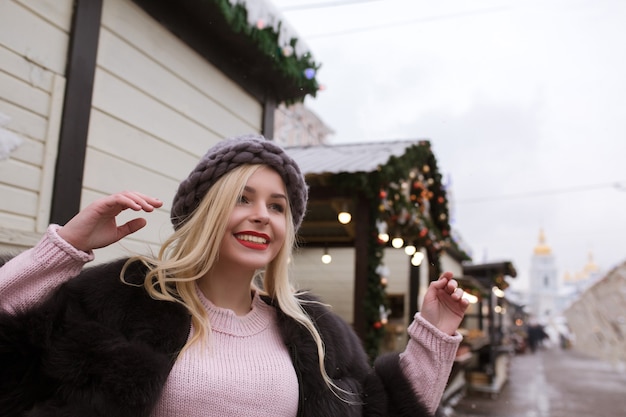 Straatportret van een vrolijke jonge vrouw met een stijlvolle gebreide muts en bontjas, poserend op de kerstmarkt. Ruimte voor tekst