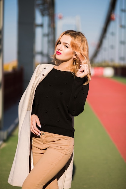 Straatportret van glamourblond model draagt stijlvolle outfit, poserend bij de brug