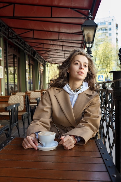 Straatportret van jonge vrouw die beige laag het drinken koffie op koffieveranda dragen met dromerige en nadenkende starende blik