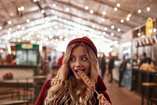 Straatvoedselcultuur. Jonge blonde vrouw in rode dop en ecobontjas die veganistische worst eet in deeg op straatmarkt.