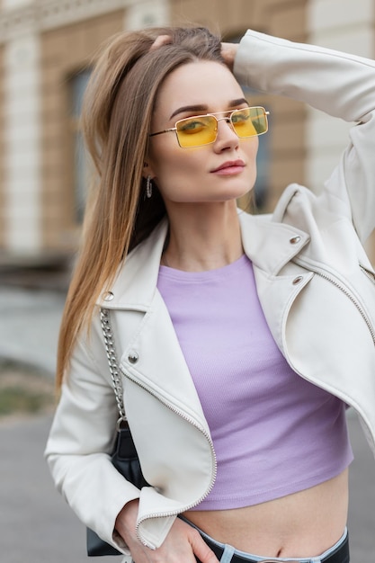 Straatvrouwenportret van een mooie modelvrouw hipster in een modieuze leren jas en paarse tshirt met een portemonnee en gele zonnebril in de stad