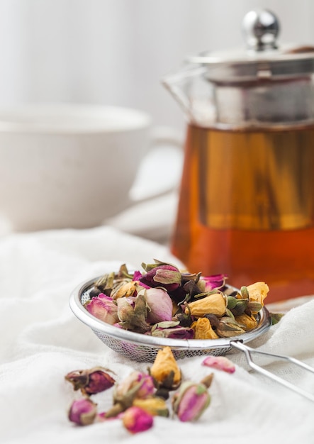 Infusore a filtro con boccioli di rosa sciolti mescola il tè su un panno bianco con teiera in vetro trasparente e tazza in ceramica bianca