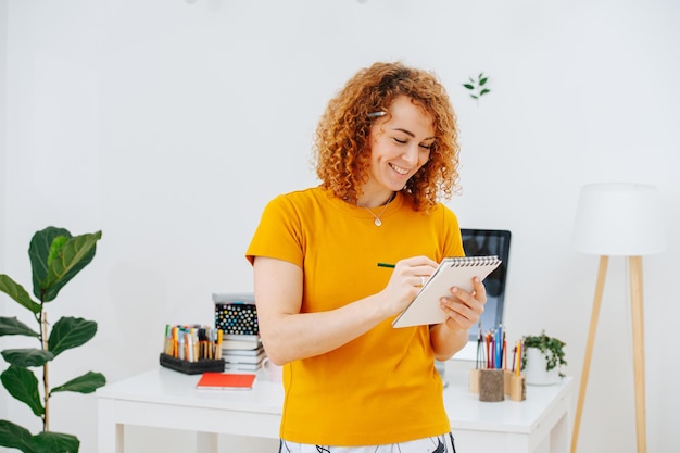 Stralende jonge vrouw met krullend gemberhaar die in haar notitieboekje schrijft