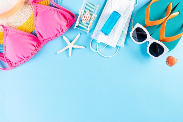 Strandhoed, bikini, zonnebril, medisch masker en handdesinfecterend middel op lichtblauwe achtergrond. Zomer.