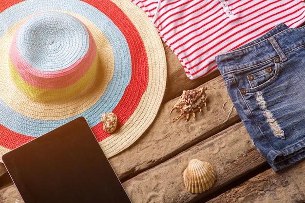 Strandhoed en zwarte tablet. Denim shorts naast schelpen. Dameskleding voor op reis. Kom en ontmoet de zomer.