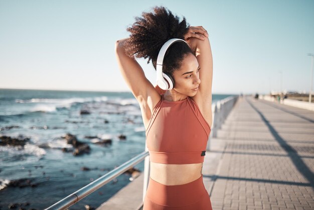 Strandkoptelefoon en vrouw die zich uitstrekt voor een fitnessoefening die hardloopt of traint voor een race of marathon Gezondheidssporten en vrouwelijke atleet die een arm opwarmt voor een cardiotraining aan de oceaan