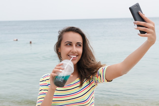 Strandvrouw die koude drank drinkt die pret heeft bij strandpartij. vrouwelijke babe in bikini die geniet van ijsthee, cola of alcoholische drank die gelukkig glimlacht. een smartphone vasthouden en selfies maken