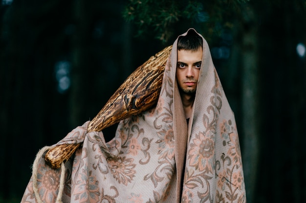 Strange man  covered with blanket standing with wooden stick in his hand in forest