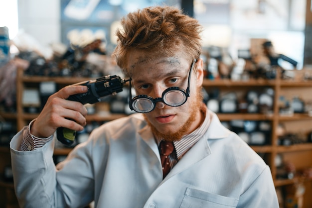 Strange scientist holds screwdriver at his temple, dangerous test in laboratory. Lab equipment, engineering workshop