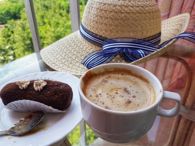 Photo straw hat and a cup of coffee with cakes on a glass table on a balcony on a summer evening