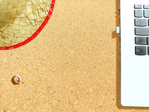 해변 모래 위의 밀짚모자와 노트북은 휴가 작업의 개념을 나타냅니다.