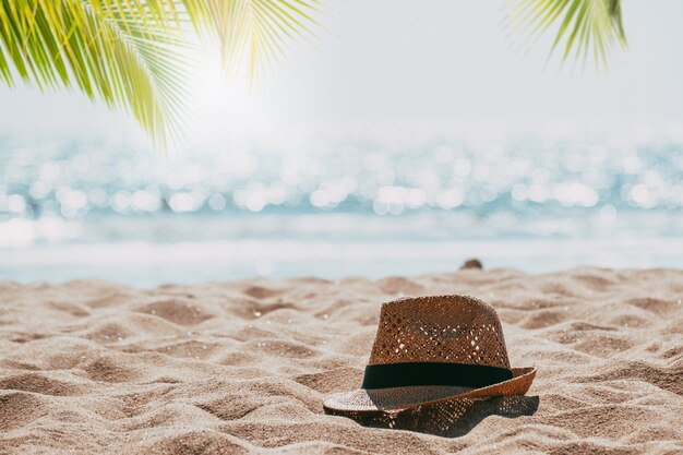 Straw hat on sand tropical beach seascape with palm tree and blur bokeh light of calm sea 