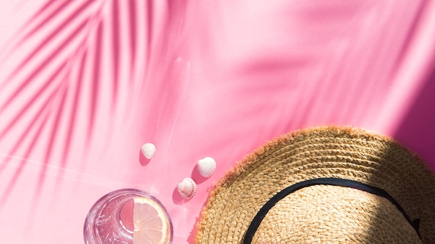 straw hat water glass and shells on pink background flat lay