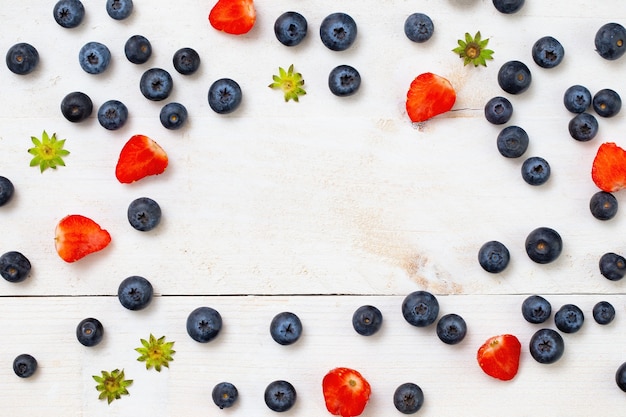 Strawberries cut in half and blueberries arranged on borders of frame with copy space in the middle on white wooden table.