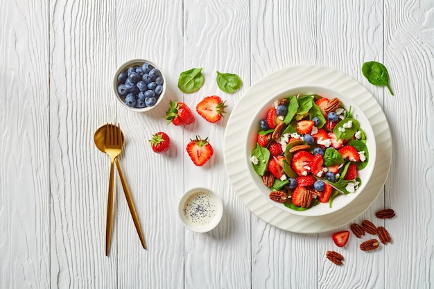 Strawberry blueberry spinach salad with pecan nuts and crumbled feta cheese in a white bowl on a wooden table with poppy seeds dressing, horizontal view from above, flat lay, free space