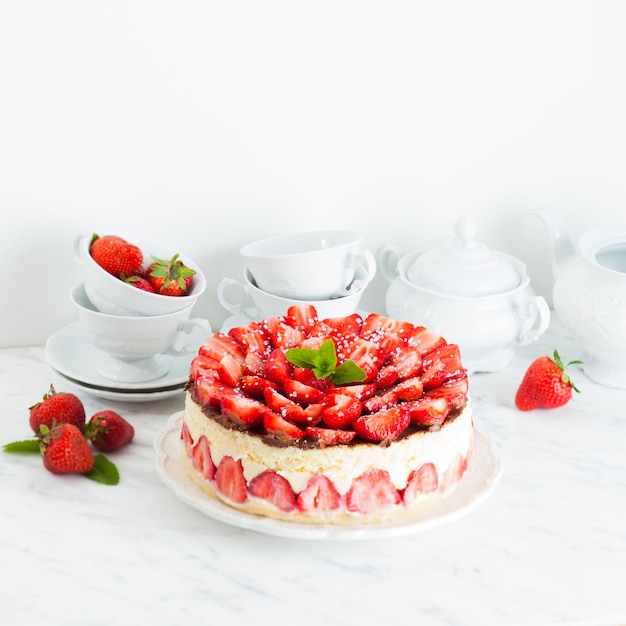 Клубничный торт Fraisier на белой тарелке и чашки для чая, ягоды и посуда на заднем плане