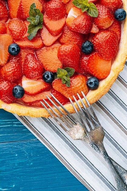 Strawberry custard pie with lime zest added to which are sliced strawberries with blueberries and fresh mint leaves and topped with lime jelly on a blue wooden table