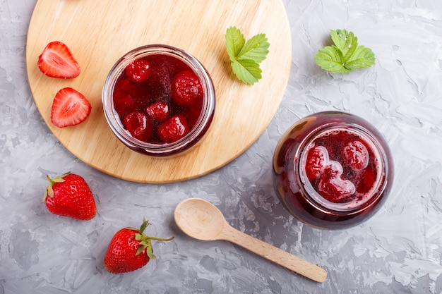 Strawberry jam in a glass jar with berries and leaves on gray concrete 